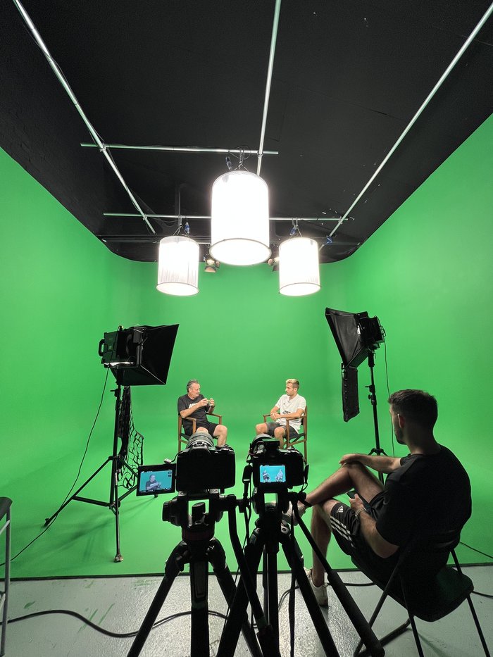 Crew filming with two cameras in front of a green screen, live monitors visible