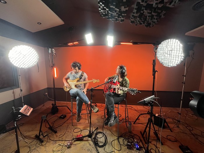 Two musicians playing guitar in a studio with an orange backdrop and studio lights